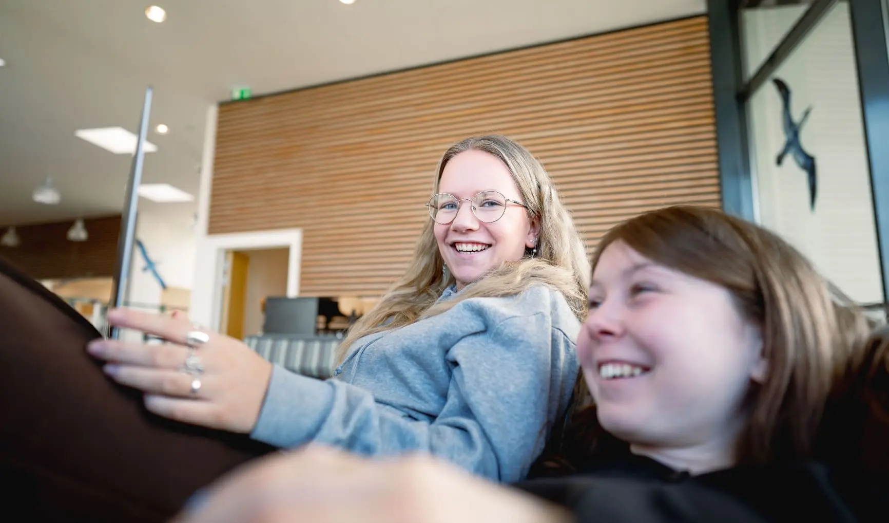 To smilende unge personer sidder i en sofa i et moderne, lyst lokale med træpaneler og glasvæg