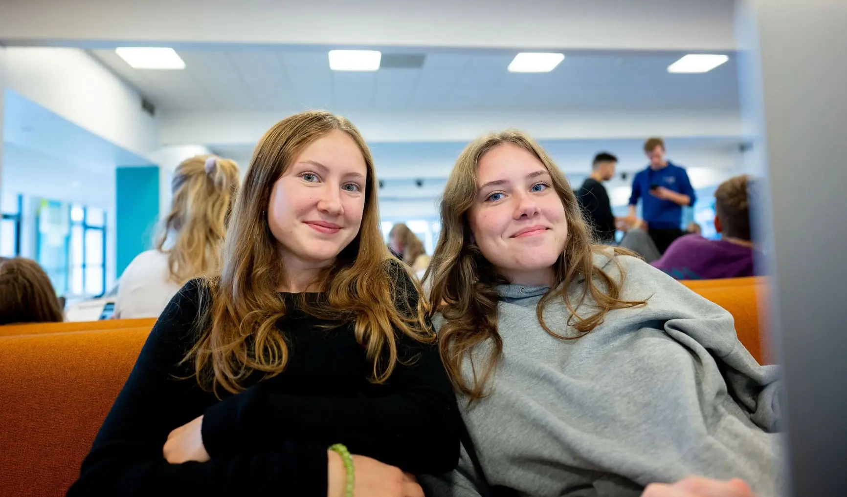 To unge kvinder sidder og smiler i et fællesområde på en skole.