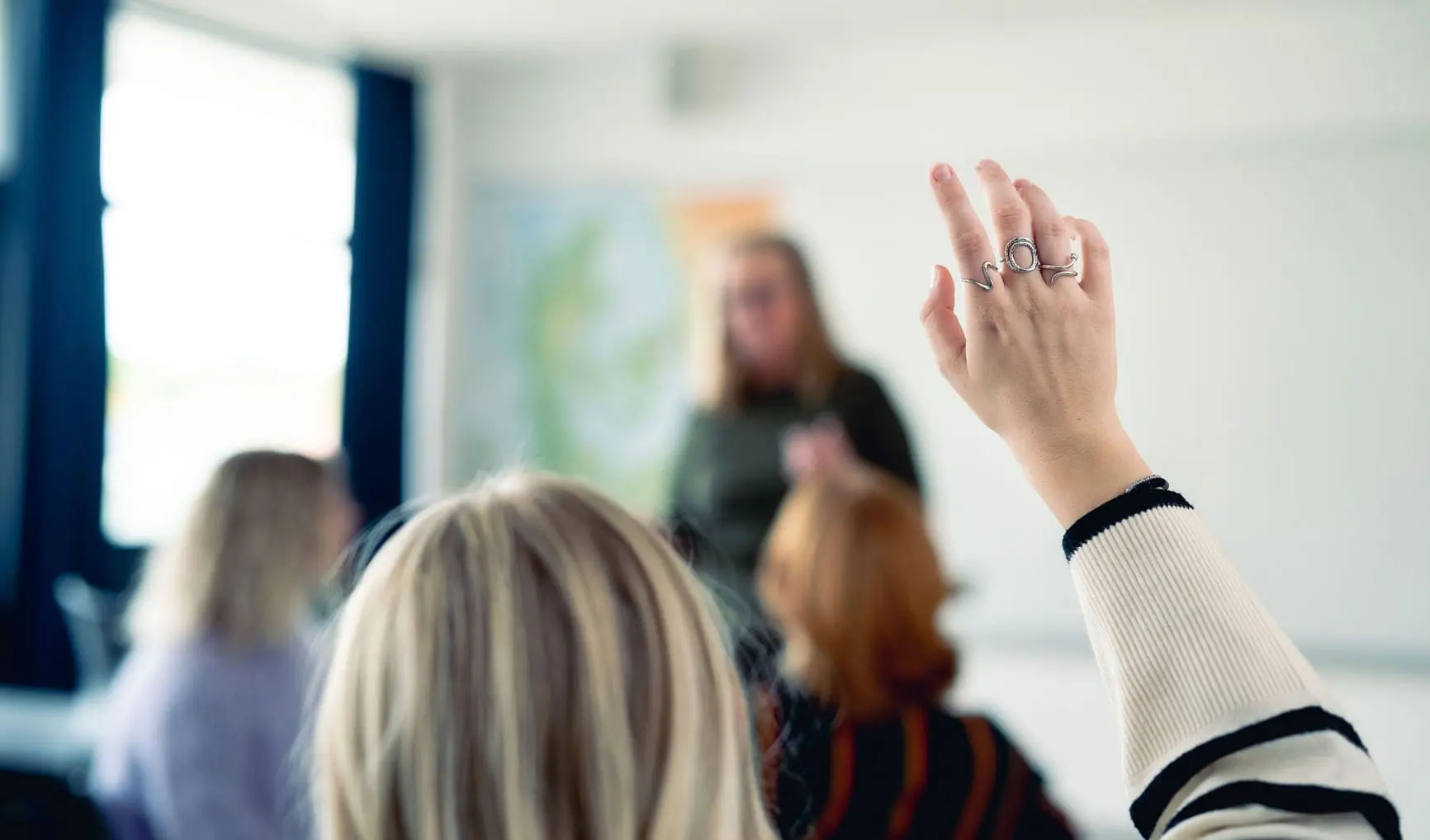 Elev rækker hånden op i et klasseværelse, hvor en lærer står foran tavlen og underviser.